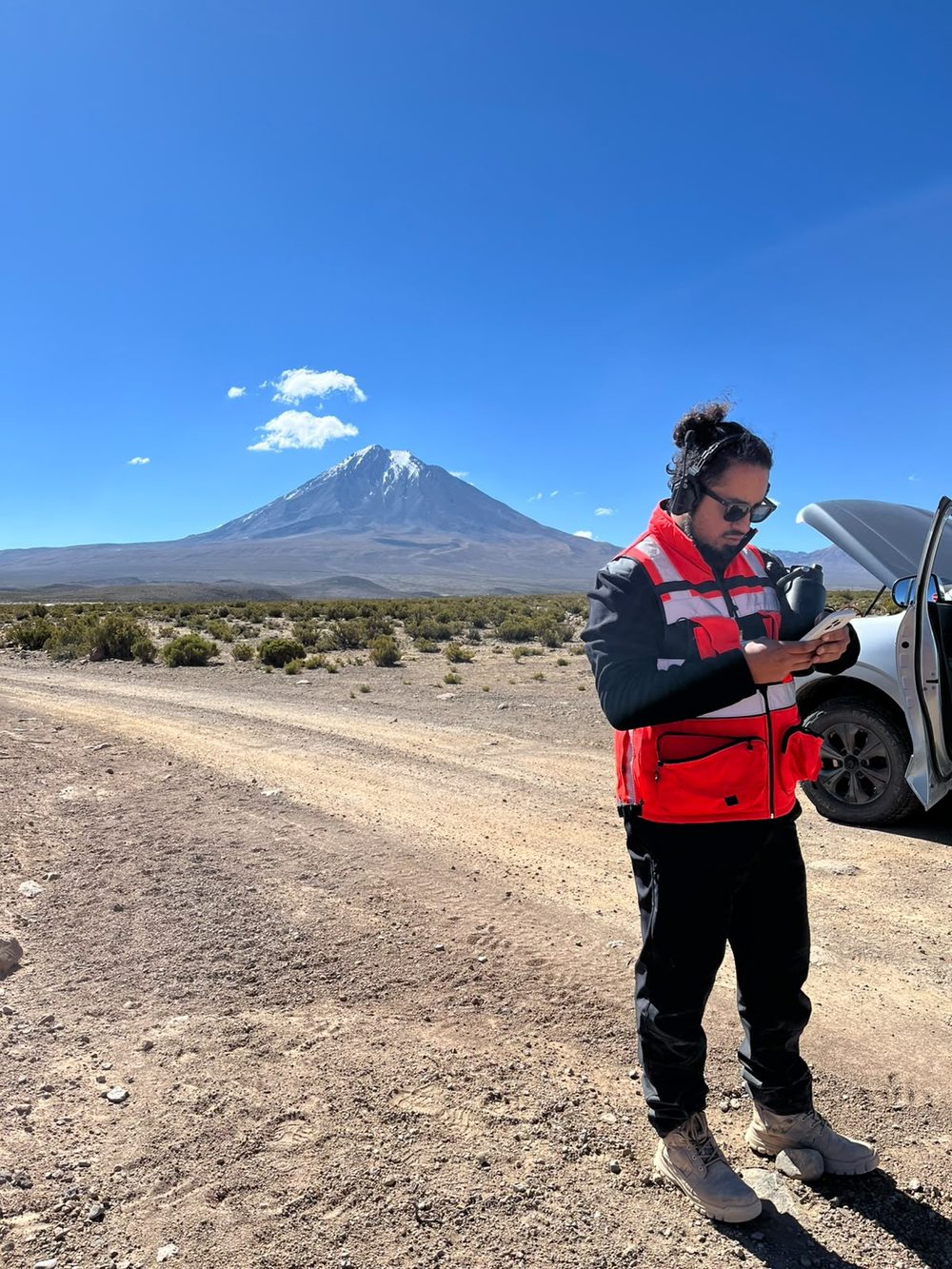 Piloto revisando la planificación del vuelo en su teléfono, junto al vehículo, con un volcán nevado al fondo